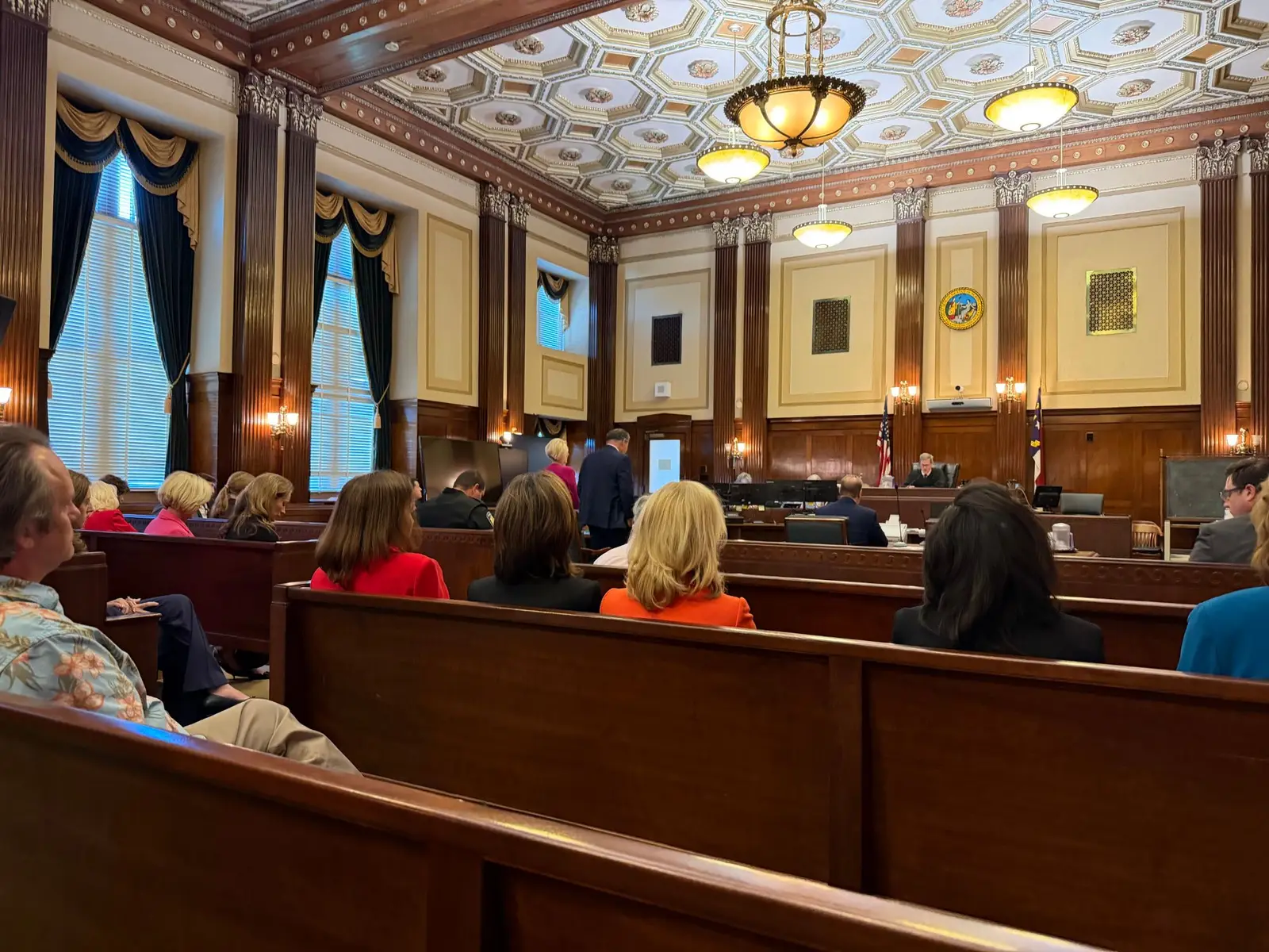 Spectators fill wooden pews in a courtroom at the Buncombe County Courthouse in Asheville, a courtroom with dark wood columns and an ornate coffered ceiling, as the judge presides from the bench.